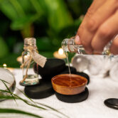 A man pours oil from a jar of essential oils standing on stones for a therapy stone and located on a terry towel next to which are transparent spheres, a twisted terry towel of white color and a sprig of lavender