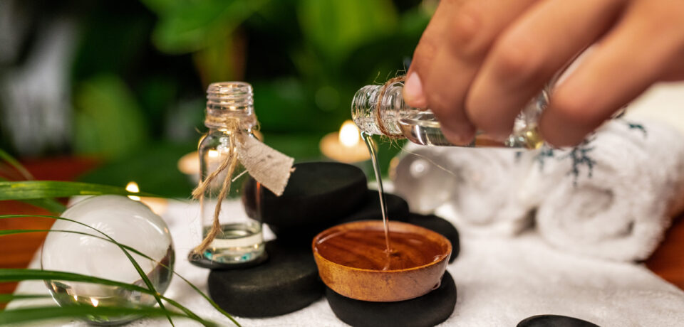 A man pours oil from a jar of essential oils standing on stones for a therapy stone and located on a terry towel next to which are transparent spheres, a twisted terry towel of white color and a sprig of lavender