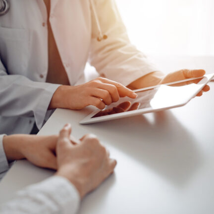 Doctor and patient sitting at the desk in clinic office. The focus is on female physician's hands using tablet computer, close up. Perfect medical service and medicine concept.