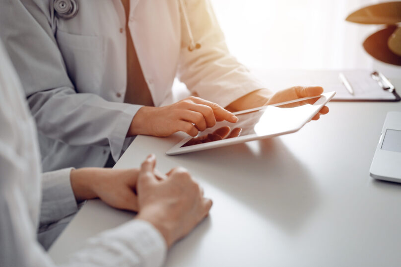 Doctor and patient sitting at the desk in clinic office. The focus is on female physician's hands using tablet computer, close up. Perfect medical service and medicine concept Doctor and patient sitting at the desk in clinic office. The focus is on female physician's hands using tablet computer, close up. Perfect medical service and medicine concept.