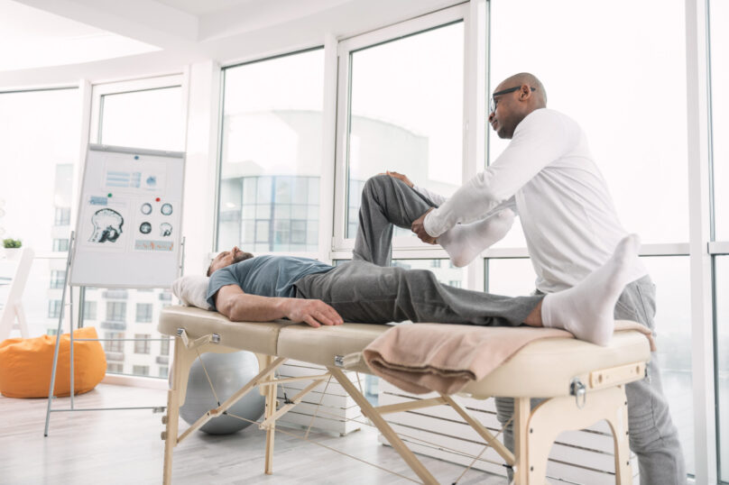 Professional physical therapist working Rehabilitation program. Professional physical therapist holding his patients leg while working according to individual rehabilitation program